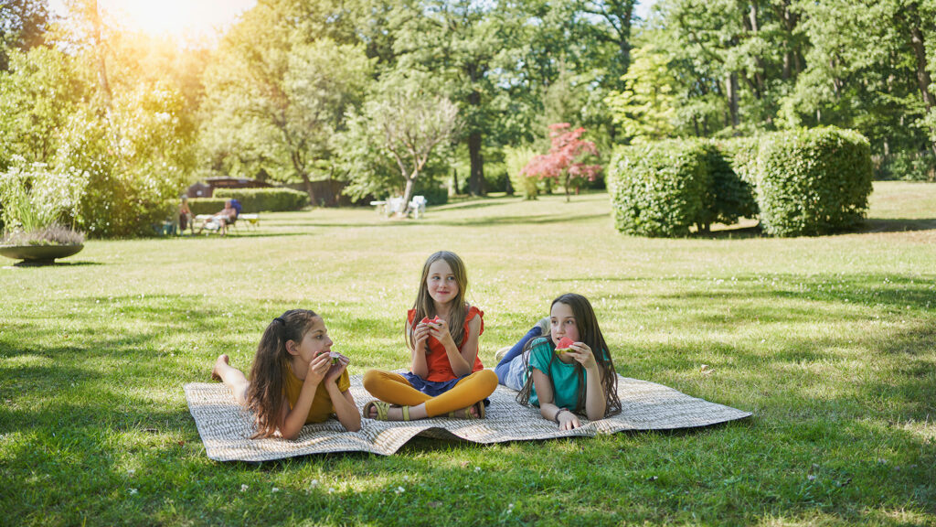 Three children are eating melon, sitting on your blanket in the grass.