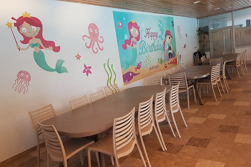 Room for the birthday party, view of tables and chairs, ‘Happy Birthday’ poster and mermaid illustration on the wall.